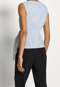 Blå och gulrandig ärmlös topp med en böjd nederkant och nyckelhålsknäppning bak, kombinerad med svarta byxor. Slät tygstruktur.