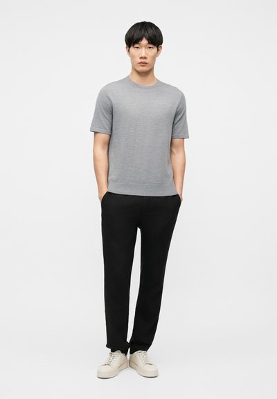 Άνδρας που στέκεται με τα χέρια στις τσέπες, φορώντας γκρι κοντομάνικο πουκάμισο, μαύρο παντελόνι και λευκά αθλητικά παπούτσια σε απλό φόντο.