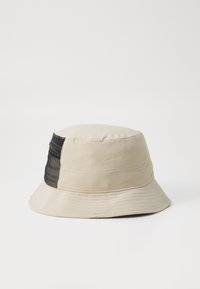 Beige bucket hat with a black mesh panel, circular crown, and wide floppy brim. Features stitched detailing and a textured finish.