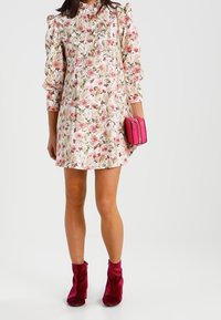 Robe courte à fleurs avec un fond blanc, imprimé de fleurs roses et vertes, manches bouffantes et yoke plissé. Associée à un petit sac à main rose et des bottines rouges.