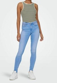 Jeans skinny blu con un lavaggio sbiadito, abbinati a un top senza maniche a righe verdi e bianche. Il modello sta in sneakers bianche su uno sfondo neutro.