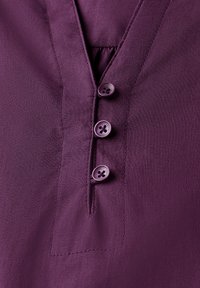Lilla bluse med V-hals, fem knaplukninger og blødt, glat stof med en subtil glans. Inkluderer en lille plissering under halsudskæringen.