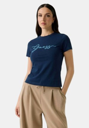 Mujer con cabello largo y oscuro, vistiendo una camiseta azul marino de Guess y pantalones beige de corte holgado, con las manos en los bolsillos.
