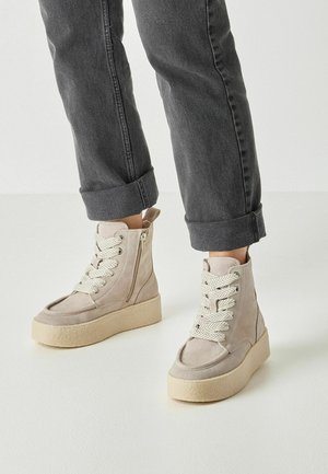Beige suede platform ankle boots with thick white laces and side zipper worn with rolled-up dark gray jeans.