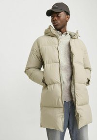 Pufferjakke i lys beige med hætte, lynlås foran og sidelommer. Båret over en grå sweater og blå jeans, med en sort kasket.