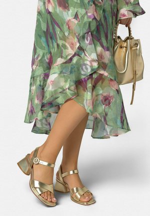 Piernas con sandalias doradas de tacón grueso y tiras, combinadas con un vestido verde floral transparente y un bolso tipo cubo dorado a juego.