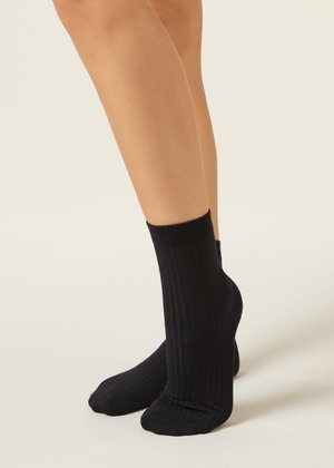 Nohy v černých žebrovaných ponožkách stojící na světle neutrálním pozadí, zobrazující texturu a střih ponožek.