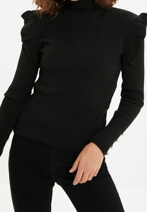 Femme portant un pull noir côtelé avec des épaules bouffantes et des boutons en perles sur les poignets des manches, associée à un pantalon noir.