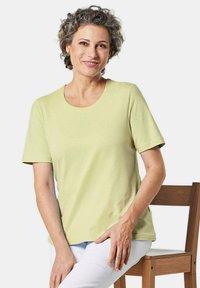 Helles grünes Baumwoll-T-Shirt mit kurzen Ärmeln, runder Ausschnitt, glatte Textur, getragen mit weißen Hosen. Model sitzt auf einem Holzstuhl.