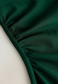 Tissu vert foncé froncé avec des coutures visibles et une surface texturée, montré en gros plan sur un fond clair.