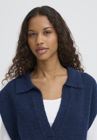 Pull tricoté oversize bleu marine avec un large col et un profond décolleté en V. Texture douce avec une finition texturée. Superposé sur une chemise blanche.