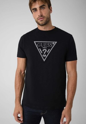 Un mannequin masculin porte un tee-shirt noir avec un logo triangulaire Guess argenté et un jean foncé sur un fond gris clair.