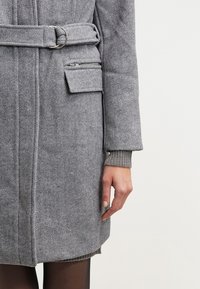 Šedý kabát se vzorem rybí kostí s páskem v pase, obsahující kapsu na zip na rukávu a žebrované manžety, vyrobený z texturované látky.