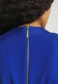 Femme aux cheveux bouclés et foncés porte un vêtement bleu avec une fermeture éclair dorée visible dans le dos au niveau du cou.