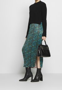 Kvinna som bär en svart tröja, svarta klackboots som når anklarna, en blå mönstrad kjol med sidospalt och håller en svart läderhandväska.