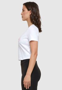Witte cropped t-shirt met korte mouwen heeft een roze grafisch ontwerp op de voorkant. Het model draagt zwarte high-waisted leggings.