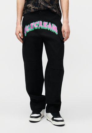 Persoon draagt zwarte cargobroek met grote kleurrijke tekst "ICECREAM" aan de voorkant en zwart-witte sneakers.