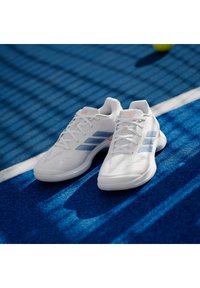 Λευκά αθλητικά παπούτσια με ανοιχτό μπλε ρίγες, υφασμένα επάνω μέρη και λευκές σόλες. Διαθέτουν κορδόνια και μια μικρή πορτοκαλί λεπτομέρεια στη γλώσσα.