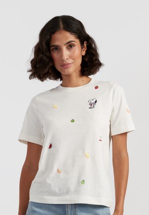 Donna con capelli scuri e ricci che indossa una maglietta bianca con Snoopy ricamato e vari piccoli disegni di frutta, abbinata a jeans azzurri.