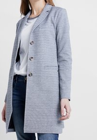 Blazer gris texturé à simple boutonnage, avec trois boutons, revers crantés et poches latérales, porté sur un haut blanc et un jean bleu.