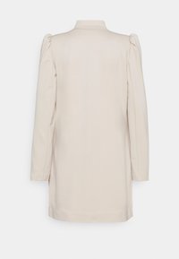 Blazer long de couleur crème avec manches longues et légères fronces aux épaules, vu de dos sur un fond uni.