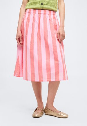 Žena v sukni s ružovými a koralovými vertikálnymi pruhmi po kolená, zelenou pletenou krátkou vestou a zlatými balerínami, stojaca na jednofarebnom pozadí.