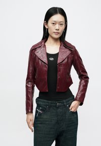 Fiatal nő egyenes fekete hajjal bordó bőr vékony dzsekit, fekete atléta felsőt és sötét derékig érő farmert visel egyszínű háttér előtt.