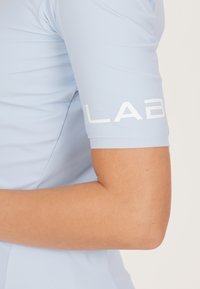 Lichtblauw sportshirt met korte mouwen. Heeft een gladde textuur en een wit logo op de arm. Fitted ontwerp dat de arm accentueert.