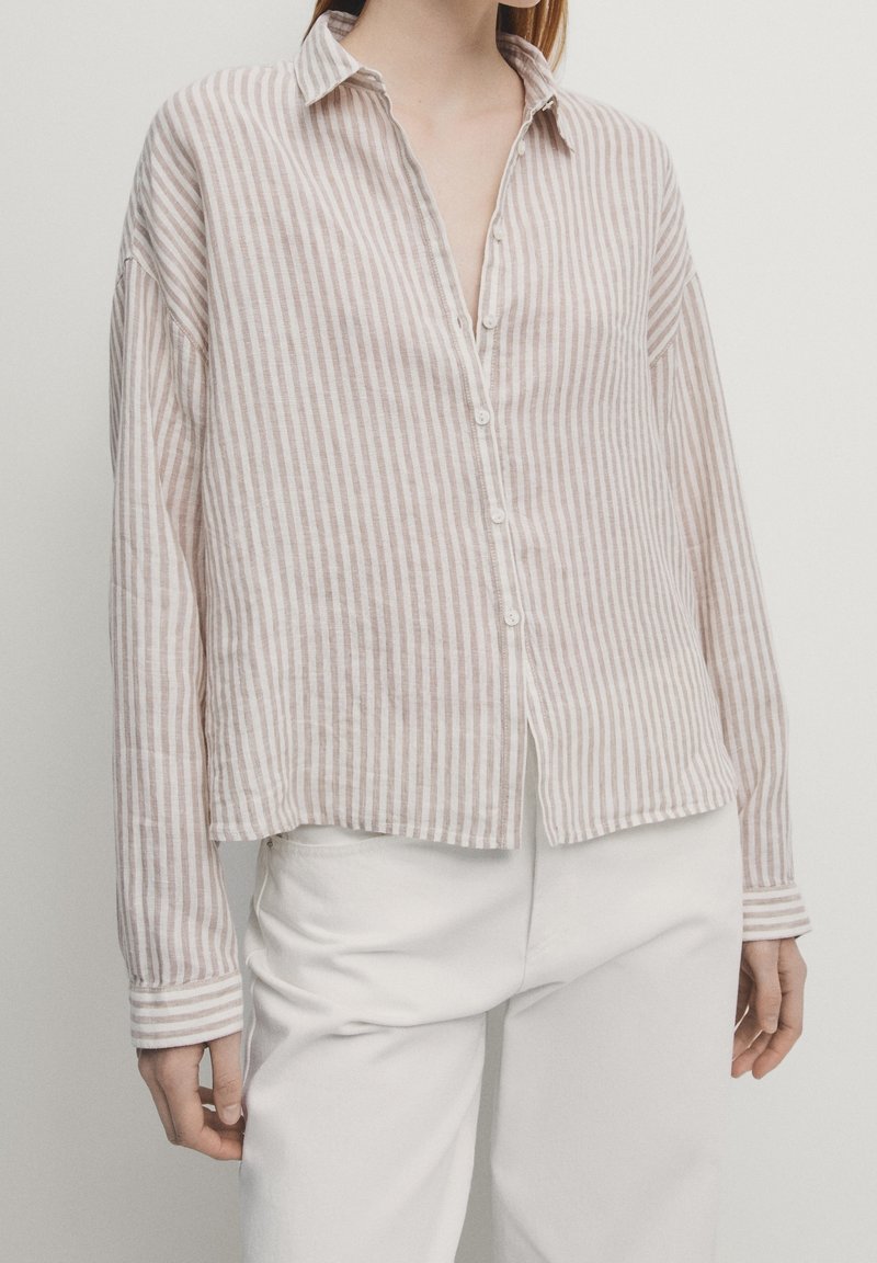 Personne portant une chemise à manches longues à rayures verticales beige et blanches avec des boutons, et un pantalon blanc, debout devant un fond uni.