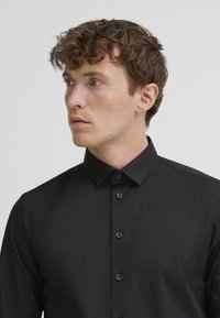 Giovane uomo con capelli castani ricci che indossa una camicia nera con bottoni, guarda leggermente verso sinistra su uno sfondo chiaro e uniforme.