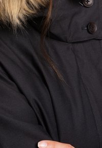 Vêtement d'extérieur noir avec une capuche bordée de fausse fourrure, deux gros boutons et une texture lisse visible dans le tissu.