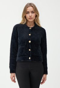 Cardigan nero e fluffy con colletto rotondo e cinque bottoni dorati davanti. Presenta maniche lunghe e una silhouette aderente.