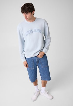 Joven con sudadera Guess Jeans azul claro, pantalones vaqueros azules hasta la rodilla, calcetines blancos y zapatillas blancas sobre fondo liso.