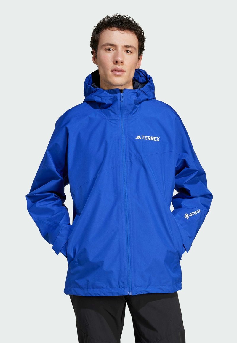 Blå vandtæt jakke med hætte, med en frontlynlås, håndlommer og et logo på brystet. Lavet af letvægts GORE-TEX materiale.