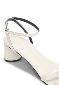 Sandalo bianco con punta aperta, tacco spesso e sottile cinturino alla caviglia con piccola fibbia dorata, mostrato su sfondo bianco.
