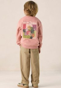 Roze sweatshirt met grafisch ontwerp op de achterkant, featuring abstracte tekeningen, gecombineerd met beige broek. Rubberzolen schoenen met groene en rode accenten.