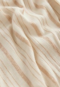 Ljust beige tyg med växelvis ljusbruna linjer, med en strukturerad väv. Materialet ser mjukt och något skrynkligt ut.