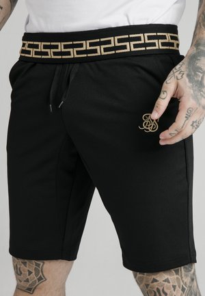 Schwarze Shorts mit beigem, geometrisch gemustertem Bund und kleinem, besticktem Logo am linken Oberschenkel, getragen von einer tätowierten Person mit weißem Hemd.