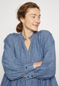 Vestido de camisa em chambray azul com colarinho volúvel, mangas longas e bufantes, e punhos com botões, apresentando uma textura suave e um ajuste descontraído.