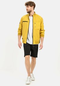 Gelbe Bomberjacke mit einem Frontreißverschluss, Brusttasche und gerippten Bündchen, kombiniert mit schwarzen Shorts und weißen Turnschuhen. Minimalistisches Design.