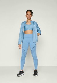 Nő magabiztosan állva, világoskék sportmelltartót, leggingset és kabátot viselve, fekete sportcipőkkel, sima háttér előtt.