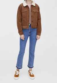 Donna che indossa una giacca in velluto a coste marrone con colletto bianco, un top rosa chiaro, jeans corti blu con orli sfrangiati e sneakers marroni con suole bianche.
