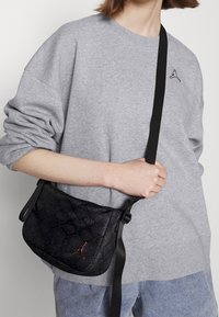 Černá kabelka přes rameno z hadí kůže s jemným embosovaným vzorem a lesklým logem. Obsahuje dlouhý nastavitelný popruh pro nošení.