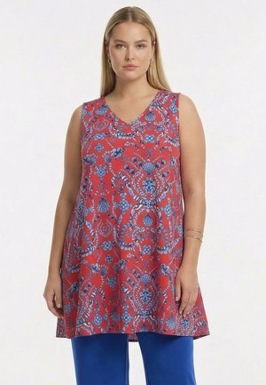 Femme portant une tunique rouge sans manches avec des motifs floraux bleus et coquillages, associée à un pantalon bleu et des bijoux simples en or.