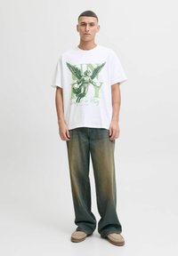 Bjela oversized majica s grafikom zelenog anđela i natpisom "JEDINSTVO", u kombinaciji s širokim izblijedjelim zelenim trapericama i svijetlosmeškim slip-on obućom.