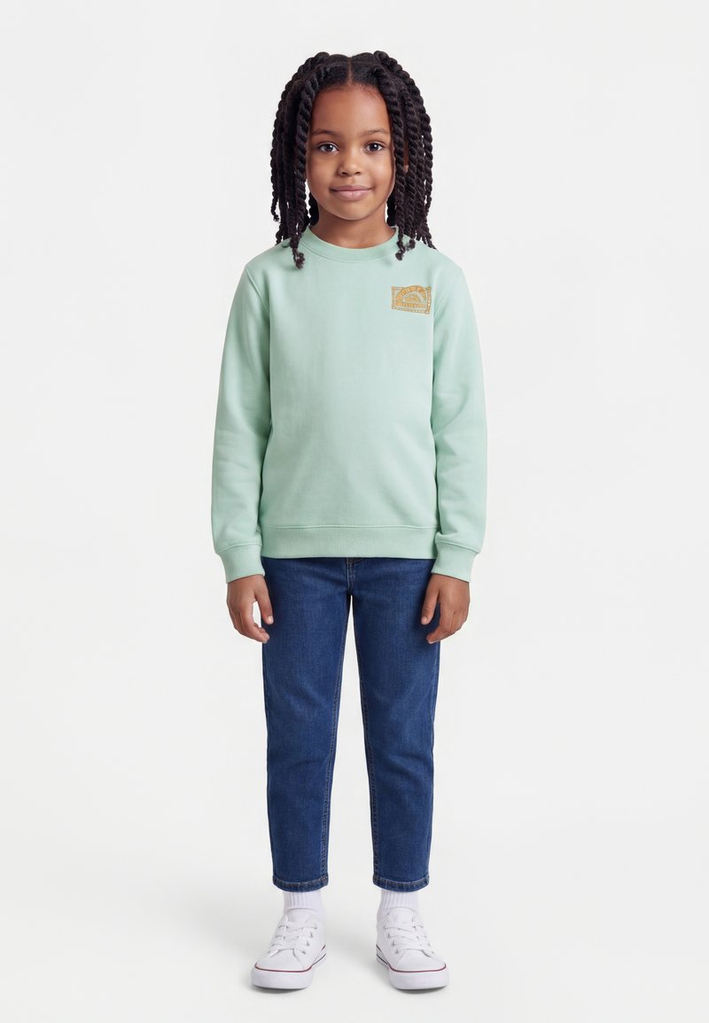 Νέα κοπέλα με μακριά στριφτά μαλλιά, φορώντας ανοιχτό πράσινο φούτερ, μπλε τζιν, λευκές κάλτσες και λευκά αθλητικά παπούτσια, στέκεται μπροστά σε λευκό φόντο.