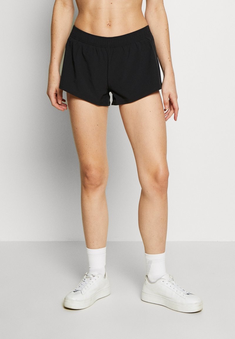 Shorts de sport noirs avec une taille élastique, présentant une coupe ample et une doublure blanche contrastante à l'intérieur de la cuisse. Associés à des baskets blanches.