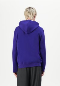 Persoon met kort blond haar, gekleed in een effen paarse hoodie en donkere broek, staande met de rug naar de camera tegen een witte achtergrond.