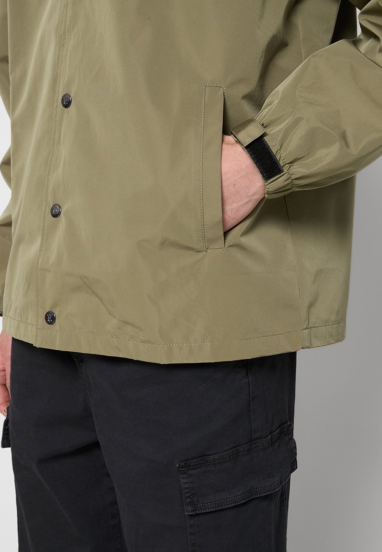 Olijfgroene, lichtgewicht jas met drukknoopsluiting, een elastische manchette en zijzakken. Gegarandeerde textuur van de stof. Zwarte broek getoond.