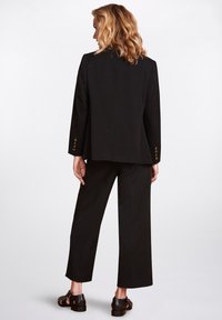 Svart skräddarsydd kostym med en strukturerad kavaj och vida byxor. Anmärkningsvärda detaljer inkluderar guldfärgade knappar på ärmarna och en mjuk tygkvalitet.